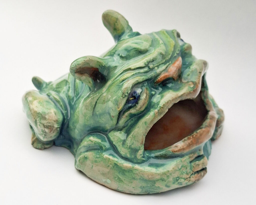 A teal blue ceramic sculpture of a squat, wrinkled creature. It has short pointy ears, expressive blue eyes, and a large, gaping mouth.
