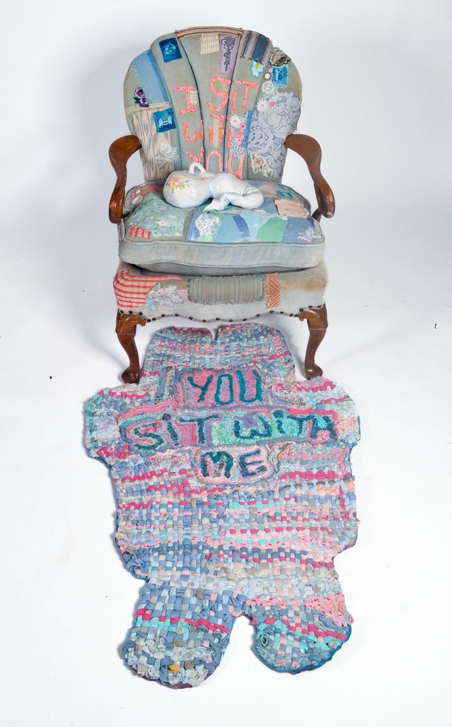 A light blue chair covered in patches and decorative stitching sits in front of a pastel coloured woven rug. The words “I sit with you” are adhered to the chair and “you sit with me” are adhered to the rug.