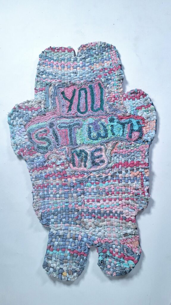 A rug woven with pastel pink, teal, blue, and purple fabric. The rug is shaped like the artist's mother's wheelchair and features the words "you sit with me" in its centre.