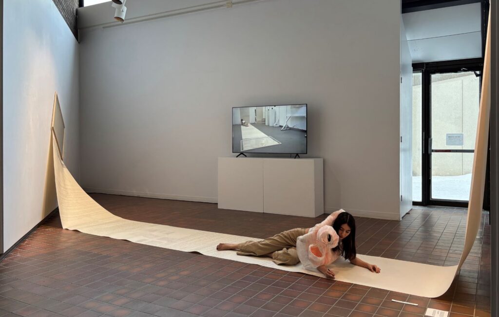 The artist is sprawled across a long tapestry extended across the room, anchored to the walls. She recounts the caretaking of her sister through handwritten testimony while adorned in a pink sculptural garment.