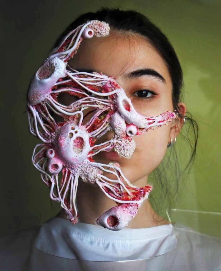 A portrait of a woman against a bright, green background. Her face is obscured by a large pink tumor, she dons a hospital gown and radiation shield.