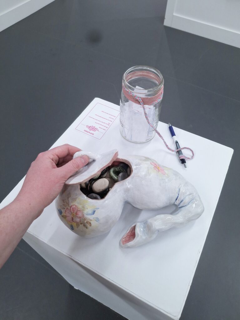 A white ceramic vessel in the shape of a gallbladder sits on a plinth. A hand lifts its lid to reveal it is filled with small stones. Beside the vessel is a jar filled with slips of paper.