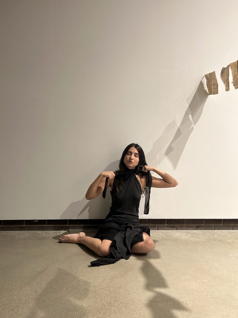 The artist sits against a white wall, legs bent, strangling herself with a piece of black sheer fabric, eyes closed, shadows of hanging paper intersect with the figure.
