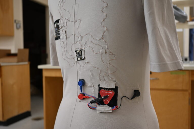 Prototype smart shirt on a dress form, with copper conductive thread traces connecting multiple EMG sensors and a microcontroller module.