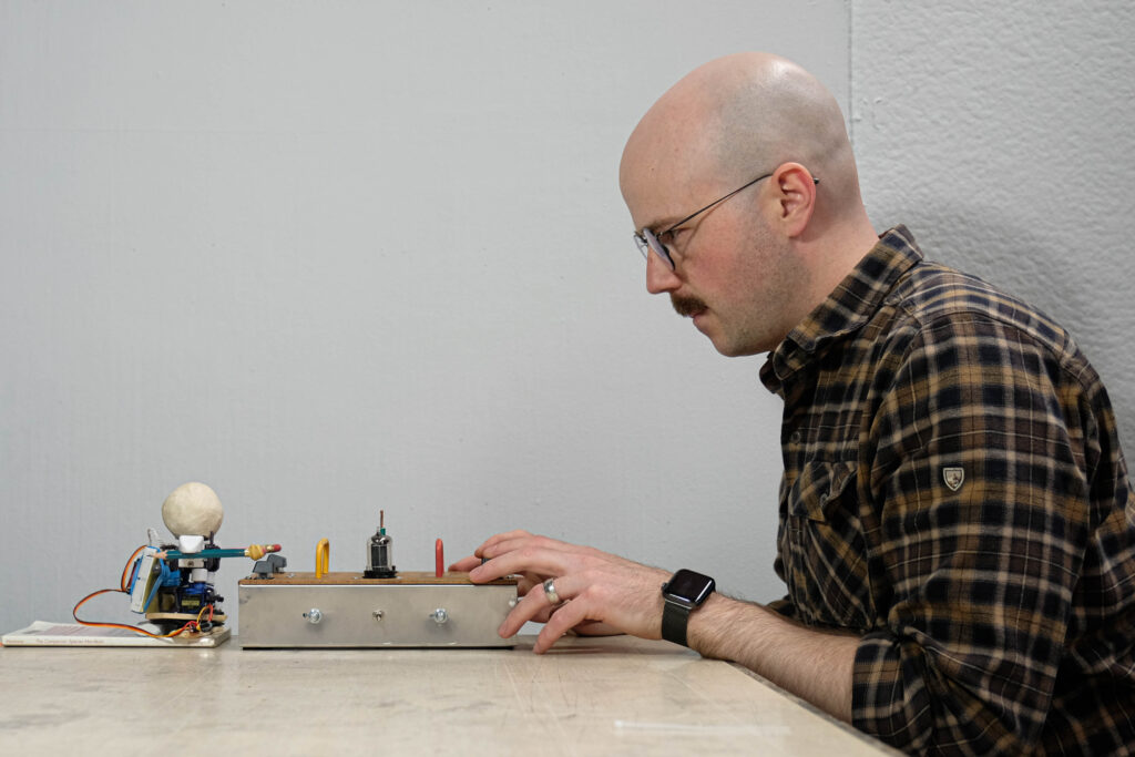 The artist sits across a table from a miniature robot, he looks at his foe intensely as they both operate switches on a panel